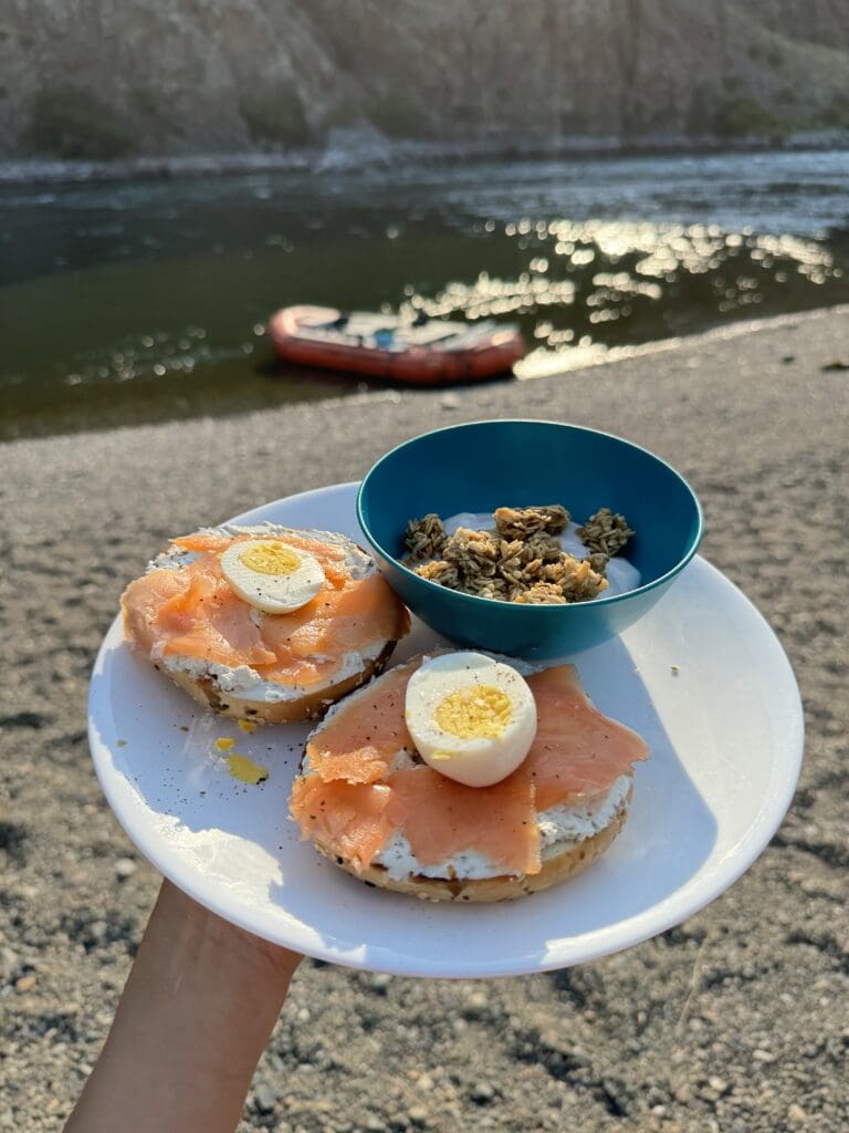 Plate with bagel topped with cream cheese, lox and hard boiled eggs with a bowl of yogurt topped with granola. The background is Hells Canyon river with a rafting boat tied up on shore.