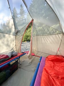 Interior view of a large camping tent with two cots and sleeping bags, looking out through the open door at a rocky riverbar and forest in morning light.