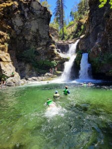 Waterfall with three separate falls and a large pool of water with a few people swimming while others sit on the rocks surrounding the pool.