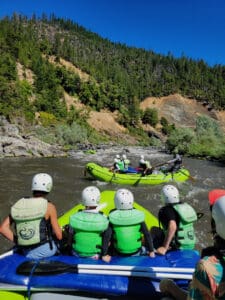 Two boats full of rafters look upstream as another boat navigates whitewater rapids. The surrounding banks are full of green trees and rocks.