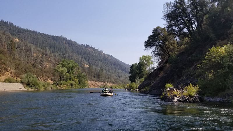 A few people on a rafting boat floating down a calm river surrounded by green trees.