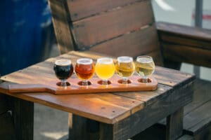 A flight of five beer beer classes with beers of various colors sitting on a wooden table next to a wooden chair.