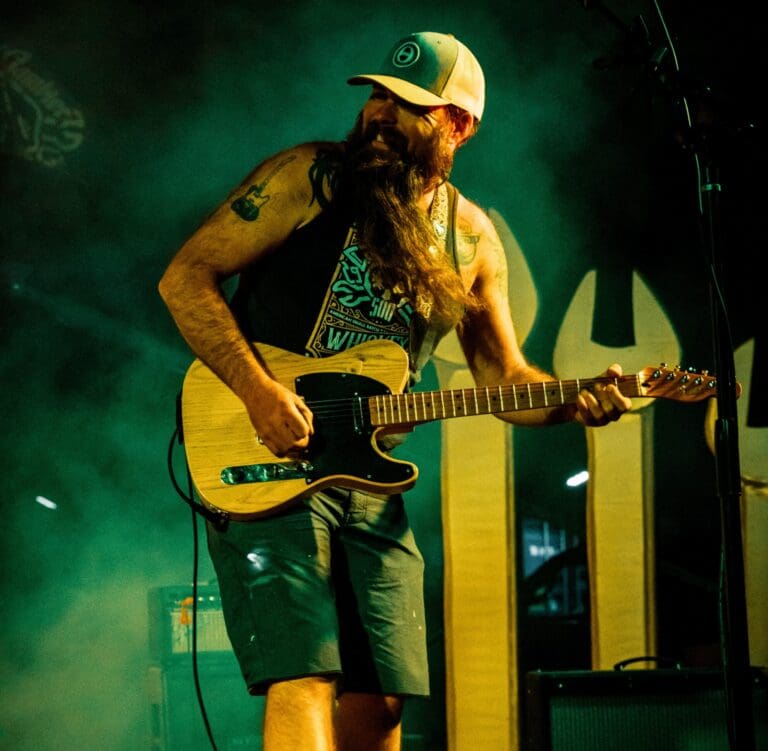 A man with a long beard playing the guitar on stage. He is wearing a hat, a tank top and shorts and has a tattoo of a guitar on his upper arm.