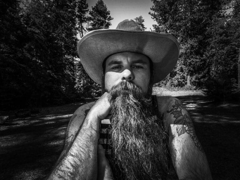 A black and white photo of a man with a long beard and a cowboy hat on. He has tattoos on his arms and is staring directly into the camera.