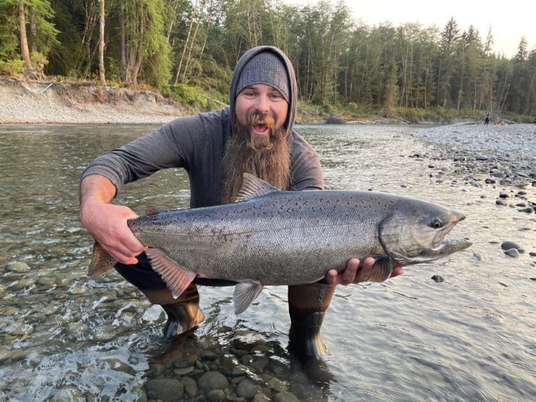 A man with a beard kneeling in a shallow calm river holding a large fish. There are green trees and small rocks lining the shores of the river.