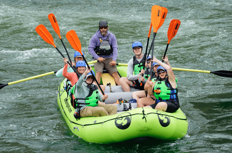 A group of people in an inflatable rafting boat, wearing lifejackets and helmets, raising their paddles in the air with big smiles. A guide sits in the back of the boat holding two large paddles. The river around them has some small white water waves.