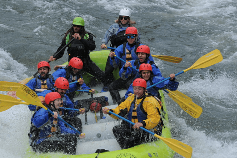 A rafting boat full of people holding paddles with a helmet and lifejackets. They are navigating a whitewater rapid and most are getting splashed by the water.