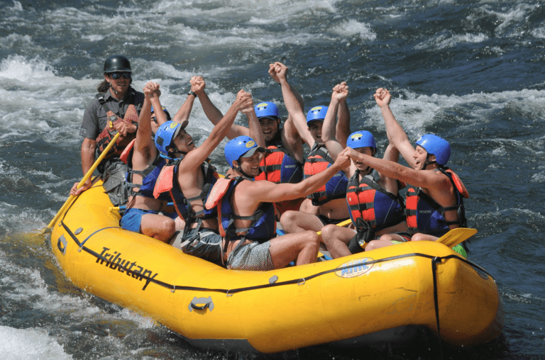 A group of people on a yellow inflatable rafting boat with one guide in the back holding a paddle. The people have their hands in the air, holding hands. There is whitewater around them.