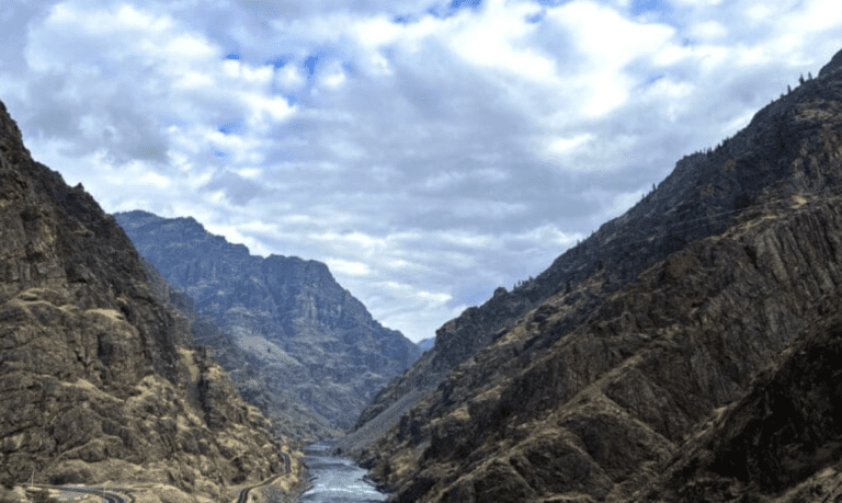 A small river in a deep canyon surrounded by huge rock cliffs of all sides. The sky is cloudy with small patches of blue sky.