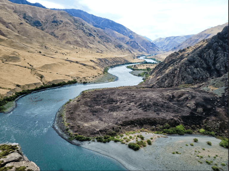 Overhead view of a winding river through a large canyon. Green shrubs line the shores with a brown grassy hill on one side and dark rocks on the other.