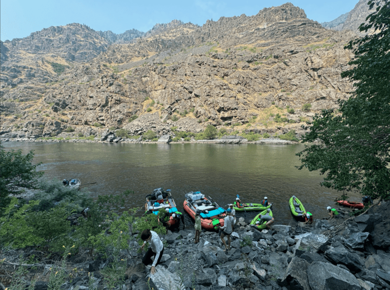 A few inflatable rafting boats and kayaks tied on the shore of a calm river. There are people in and around the boats. The river is surrounded by large rocks.