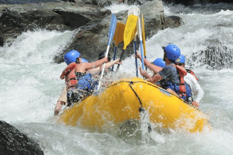 A group of adults in a yellow inflatable rafting boat navigating a large whitewater rapid. There are several large rocks in the river. The rafters are holding their paddles to the center of the boat, doing a "paddle high five"
