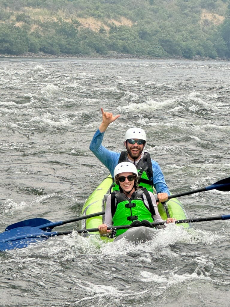 Two people in a kayak navigating white water in Hells Gorge. They are wearing helmets and lifejackets, holding paddles while smiling.