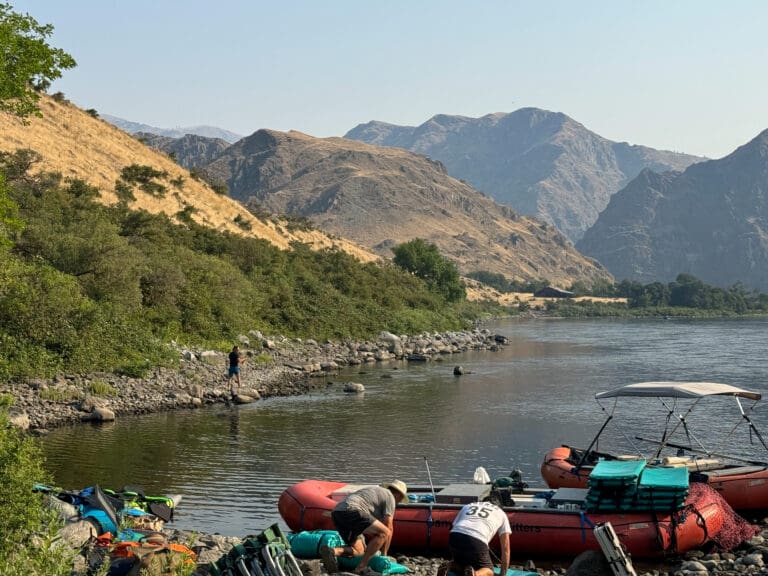 Who men on their hands and knees preparing gear and loading it onto inflatable rafting boats. Various gear on shore and on the boats. Another person is walking along the rocky shore next to the boats.