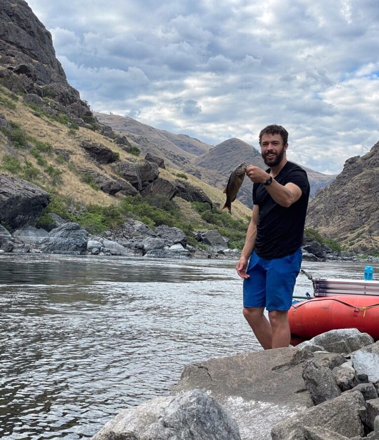 Man standing next to a river holding up a fish with a big smile. There is a rafting boat behind him and large rocks around the shore.