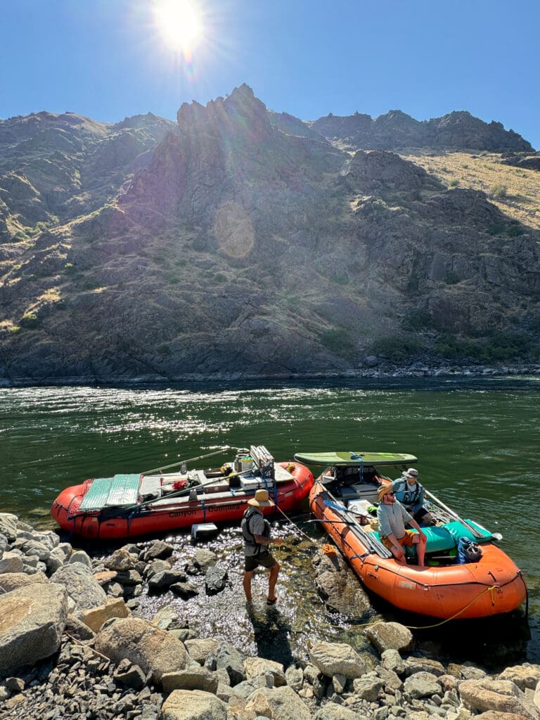Sun shining down on large rock face and river. Two inflatable rafting boats loaded with gear including a paddle board and paddles. Two men are in one of the boats and another stands in the water next to them.