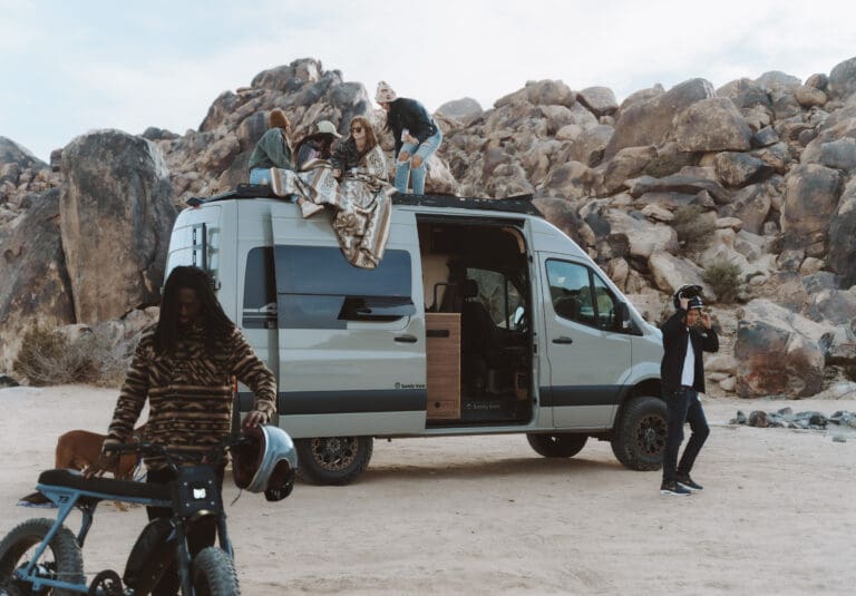 People hanging out around an adventure van. Some people on roof under a blanket, two men putting on helmets, getting ready to ride an electric bicycle.