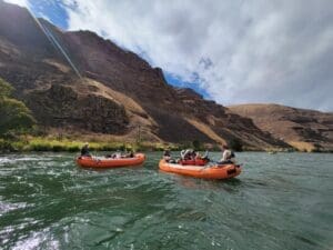 Two orange inflatable rafts with passengers floating on a wide green river beneath towering dry canyon walls under a partly cloudy sky.