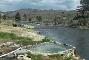 A natural stone-rimmed hot spring pool in the foreground with a rafting group and green rafts pulled ashore along a river valley backed by sage-covered hills.