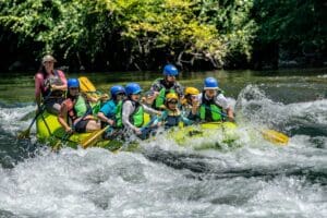A group of adults and children in blue helmets and green life jackets paddling a yellow Raft California raft through splashing whitewater, guided by a smiling female guide.