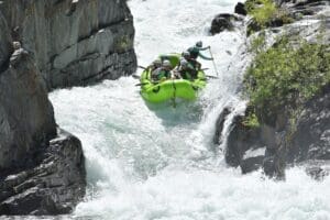 Six rafters in a green inflatable raft perched at the brink of a powerful waterfall drop between sheer granite walls, with whitewater crashing below.