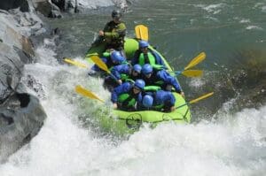 Seven rafters in matching blue jackets and blue helmets paddling a green raft through a violent whitewater rapid between boulders.