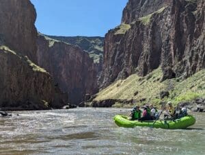 A green raft with five passengers floating toward a narrow gap between massive sheer canyon walls under a clear blue sky.