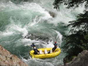 Aerial view of three rafters in a yellow inflatable raft navigating turbulent green whitewater rapids beside a rocky cliff and pine trees.