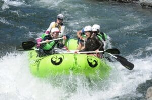 Five rafters in helmets and life jackets paddling a bright green Maravia raft through crashing whitewater, with one rider reacting in wide-eyed surprise.