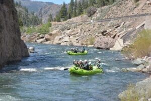 Two green rafts with helmeted paddlers floating down a river canyon with rocky cliffs, pine forests, and a railroad track visible on the hillside.