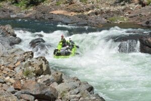 Four rafters in a green inflatable raft navigating a churning rapid between large boulders in a rocky river canyon.