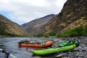 An orange Canyon Outfitters gear raft and a green Tomcat Tandem inflatable kayak pulled ashore on a rocky riverbank with sweeping canyon hills in the background.