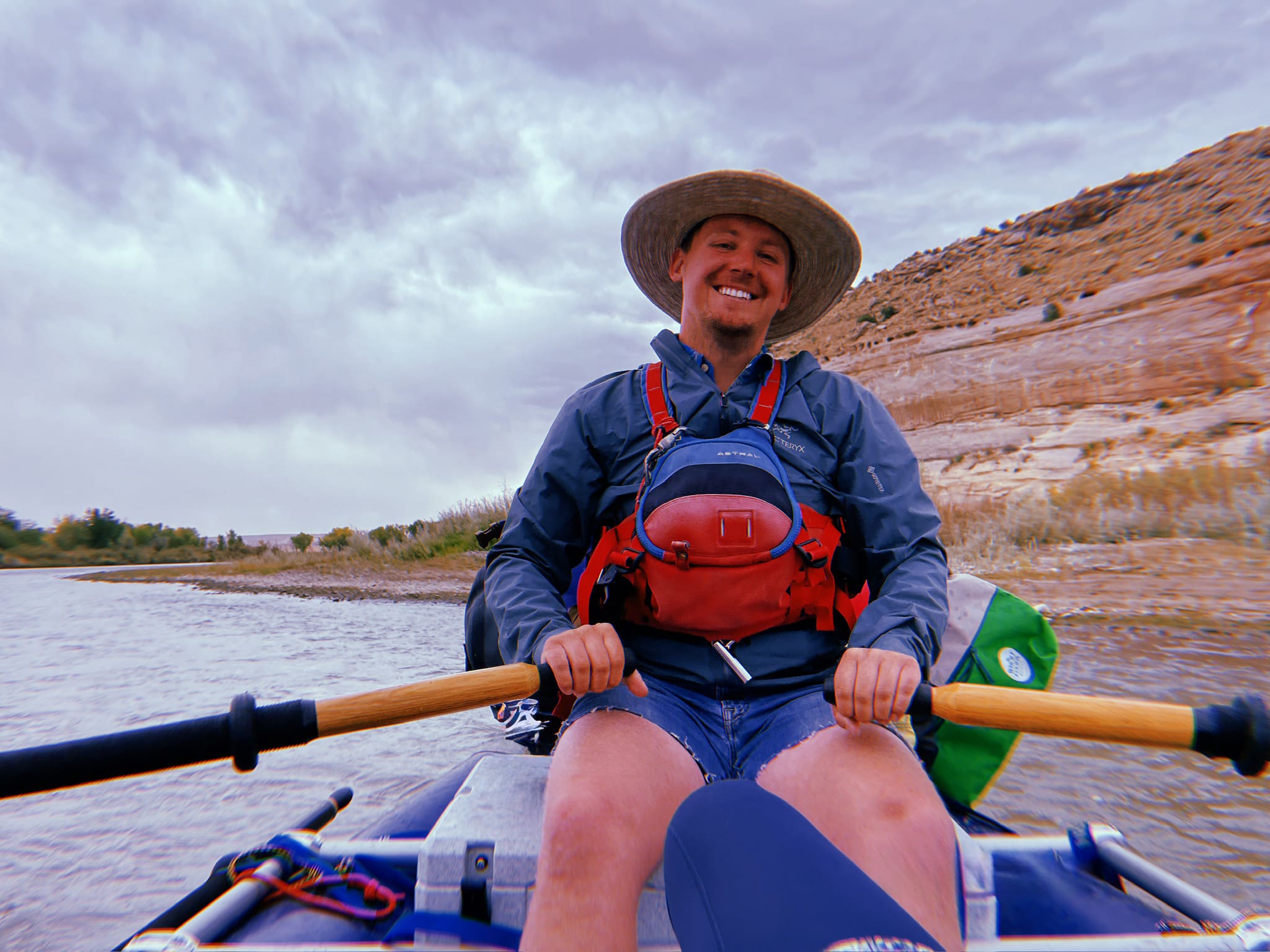 Smiling young man in a straw hat and life jacket rowing a raft on a desert river with sandstone cliffs under a cloudy sky