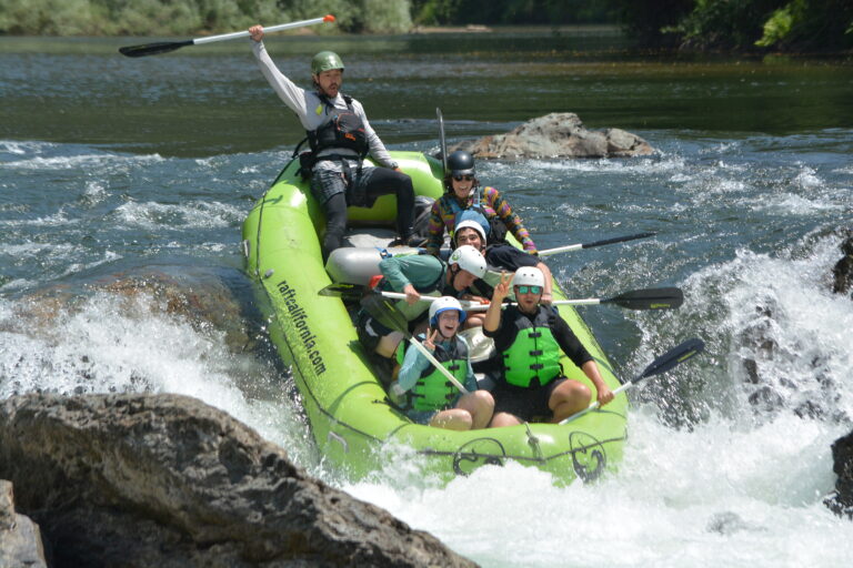 A group of six people in helmets and life jackets laughing and cheering on a bright green raft navigating whitewater rapids
