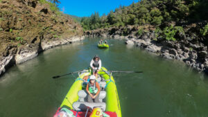 A GoPro-style shot from the bow of a Raft Oregon raft showing two rafts ahead navigating a narrow dark rock canyon corridor with conifers above